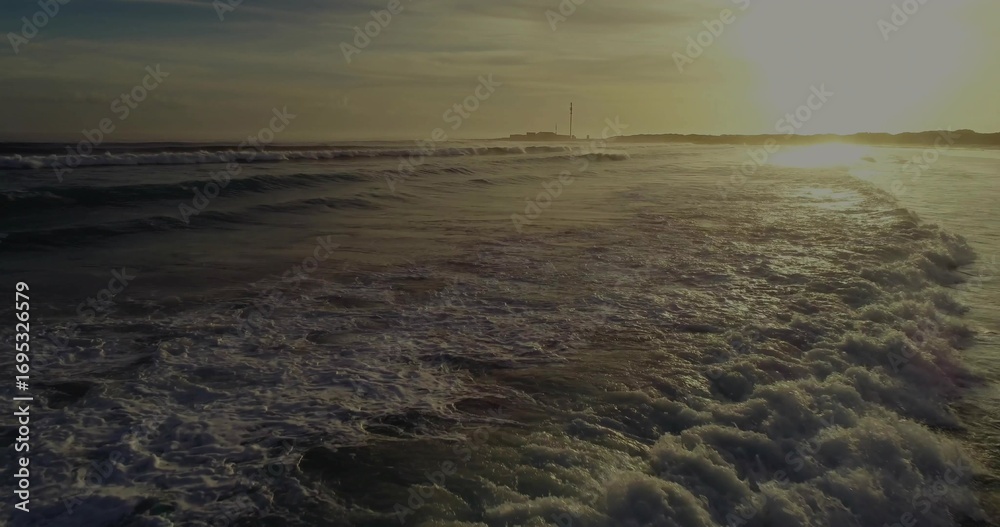 Fototapeta premium Rolling foamy ocean waves catching golden sunset light at beach viewpoint, coastal seascape