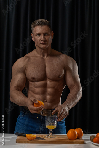 In a kitchen with dark curtains a muscular man is squeezing oranges into a glass while surrounded by fresh oranges. The focus is on his strength and commitment to healthy living.
