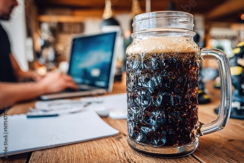Dark beer in a mason jar mug on a wooden table, alongside a laptop and paperwork
