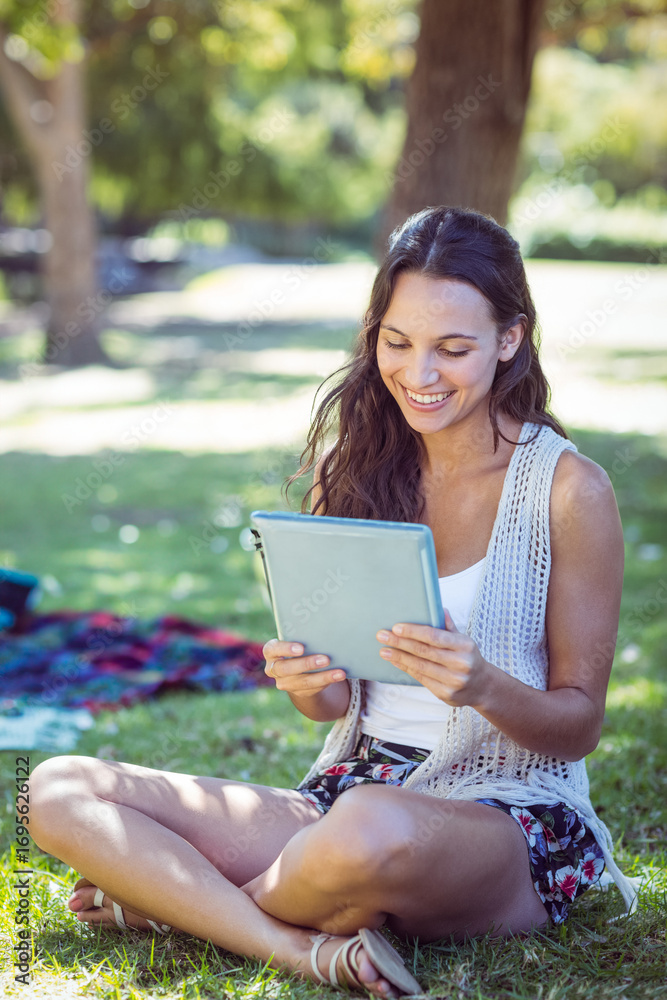 Obraz premium Woman sitting cross-legged on grass in park holding tablet near picnic blanket smiling at screen