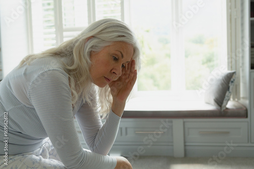 Free Download Senior woman sitting on bench under window shutters in bedroom, pressing hand to temple, copy space - GFXLoad