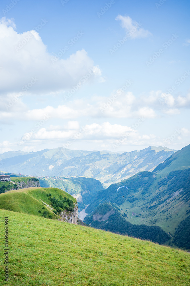 Obraz premium Beautiful landscape of a paraglider flying above a lush green valley in the Georgian mountains, with dramatic cliffs, rolling hills, and a river below under a bright blue sky in Kazbegi, Georgia