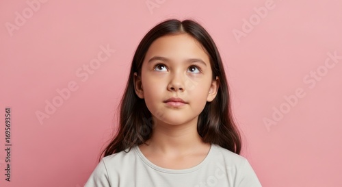 A young girl with brown hair and eyes is gazing upwards against a pink background wearing a plain white top