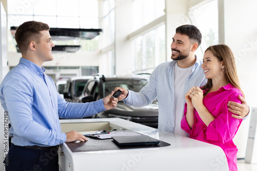 Smiling car dealer handing over keys to happy couple at dealership counter, man and woman receive vehicle keys after signing contract. Lifestyle, trust and transport