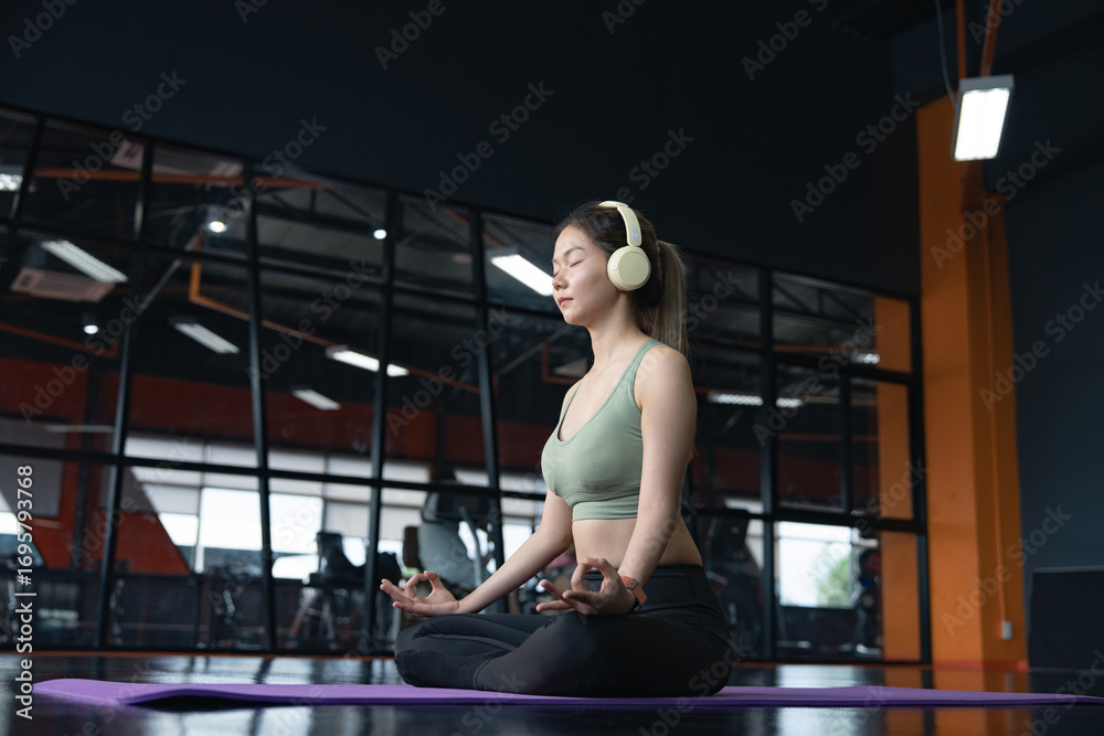 Naklejka premium Young woman sitting on the floor on mats and practicing yoga in lotus position with eyes closed in a gym.