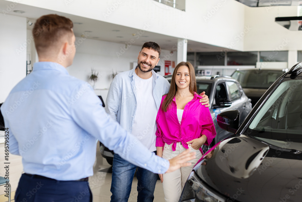 Obraz premium Smiling couple listening to car dealer while viewing vehicle in showroom. Man and woman discussing automobile options with salesman, closeup