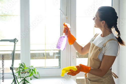 Woman in rubber gloves and apron manually washes window balcony of house with rag cleaner and mop inside interior. Restoring order and cleanliness in spring, cleaning servise