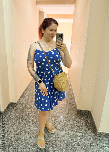 Woman in blue polka dot dress taking selfie in elevator mirror with round straw bag. Fashion, summer style, and lifestyle identity