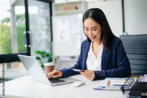 Confident Asian businesswoman smiling while using smartphone and credit card at office desk.
