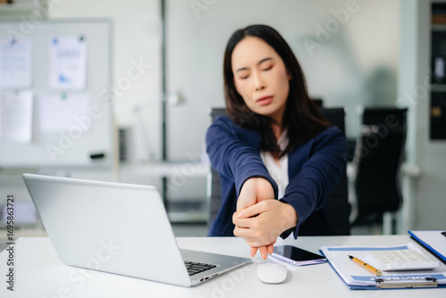Asian businesswoman stretching arms in office with tired expression, relieving muscle tension from long work hours.
