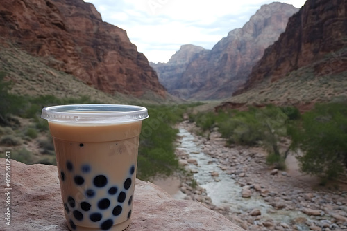 A clear plastic cup of boba milk tea with black pearls standing on a rock with a wide desert valley stretching behind 