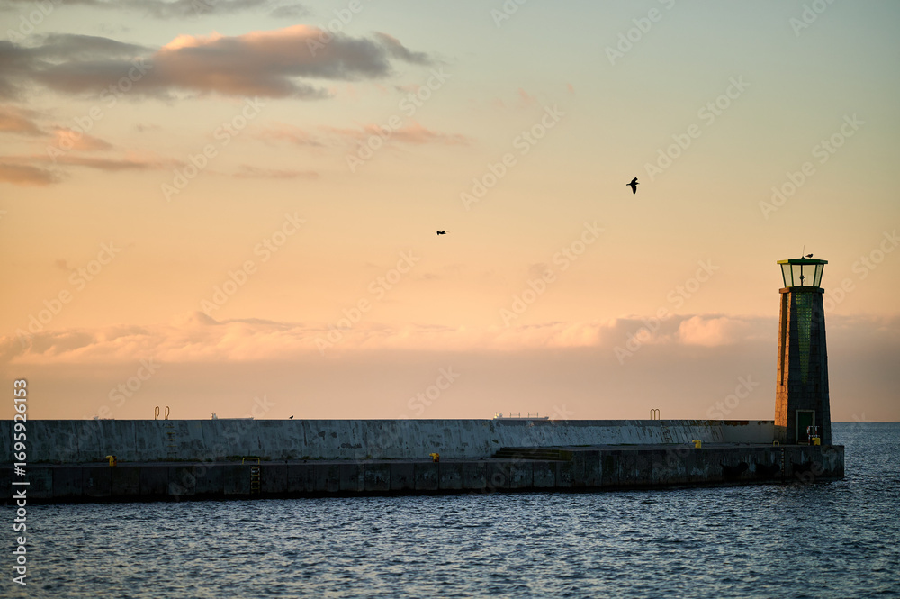 Fototapeta premium A lighthouse standing at the edge of a long pier extending into the sea, with the golden glow of the setting sun casting soft light on the water and sky.