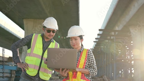 Construction Team Reviewing Blueprints: Two professional engineers, equipped with safety gear, are meticulously reviewing blueprints on a laptop at a construction site.