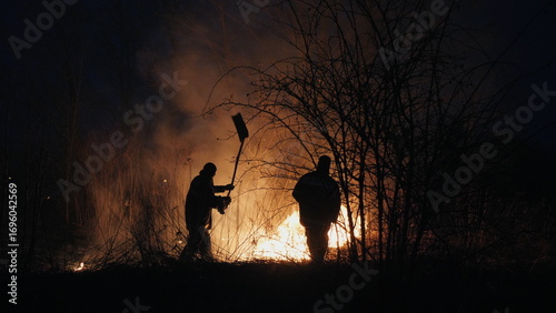 Two firefighters with fire flappers extinguish a fire in the forest at night