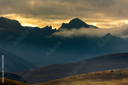 View of rugged mountain peaks pierce through a hazy sky, partially veiled by low-lying clouds, casting shadows on the golden slopes, Deosai National Park, Gilgit Baltistan, Pakistan.