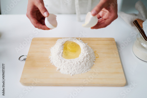 Unknown man breaking egg into mound of flour on wooden cutting board, preparing dough for baking, hands visible holding eggshells above flour and egg mixture