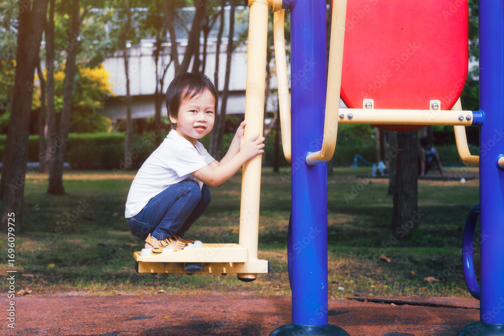 Fototapeta premium Happy Asian boy is playing on the exercise equipment in the outdoor playground, sweet smiling kid aged 3 years old is trying new things. In summer or spring.
