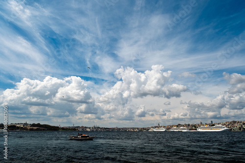 In the foreground is a small boat, behind it is a large red cargo ship. In the background is the Süleymaniye Mosque and the Istanbul skyline.