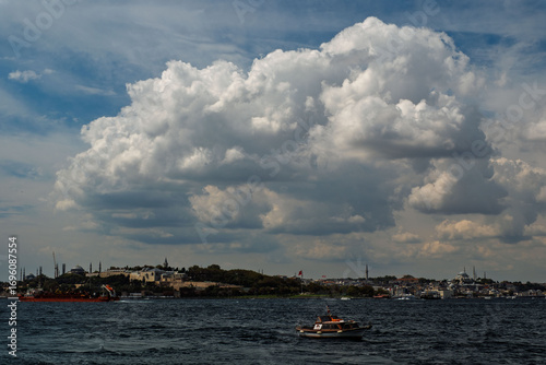 A view of the Bosphorus. Topkapi Palace and Hagia Sophia are visible on the left, and the Blue Mosque is on the right. A small boat is in the foreground, and a cloudy sky is in the background.