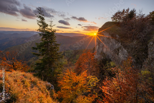 View of the sun bursting over the cliffside, casting a warm glow on the autumn trees and distant mountains, Majerova skala rock, StarÃ© Hory, BanskobystrickÃ½ kraj, Slovakia.