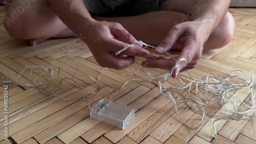 Close-up of a man's hands untangling a string of small LED fairy lights on a wooden floor. Represents home decor, DIY projects, and holiday preparation