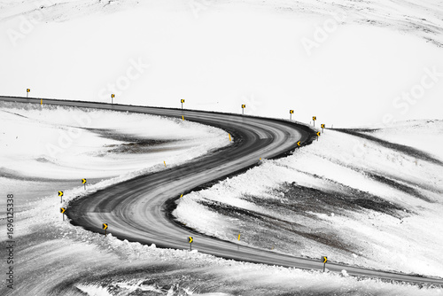 View of a winding road cuts a dark swathe through the stark white snow-covered landscape, punctuated by warning signs, in Iceland, Iceland.