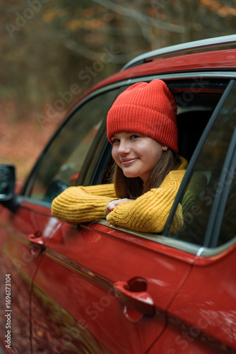 Portrait of a teenage girl in the window of a red car against the background of an autumn forest. Enjoying life.