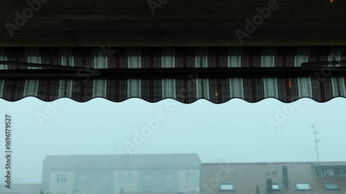 Heavy rain falling on a red and white striped awning in a gloomy urban landscape, with distant buildings obscured by fog on a dark and stormy day