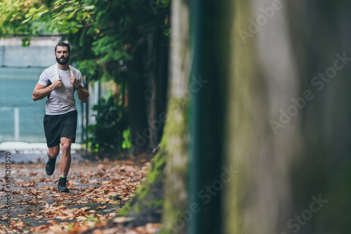 Fototapeta Naklejka Na Ścianę i Meble -  Determined young man running outside in the park. Fit boy doing exercise outdoor. Beautiful sporty male run alone. Sweaty athlete training with towel. Wellness, health, sport, fitness open air concept