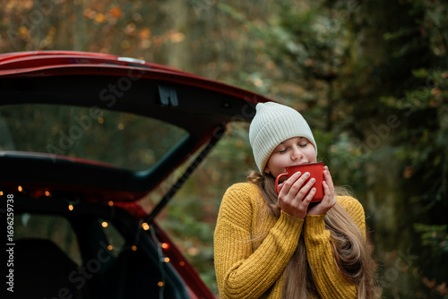 Cute teenage girl drinks hot tea from a large thermos in the trunk of a red car in an autumn forest. Picnic.