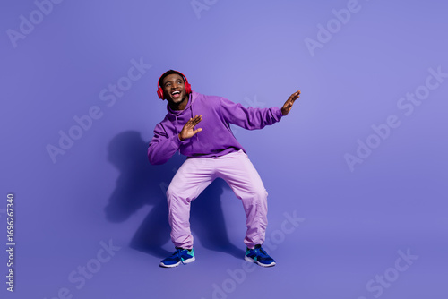 Cheerful young man in vibrant sportswear listening to music and dancing joyfully against a bold purple background