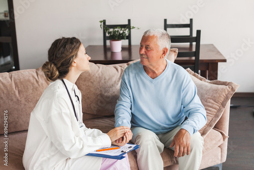 Female professional doctor showing medical test result explaining prescription using clipboard visiting senior elderly old man patient at home sitting on sofa