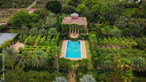 Drone video of a Spanish public garden featuring a historic ivy-covered building, elegant topiary, palm trees, and a pool surrounded by lush greenery.