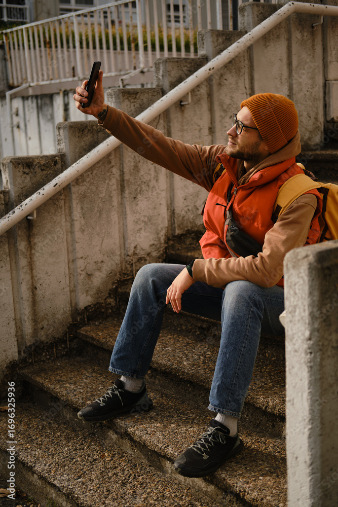 Fototapeta premium Man sitting on stairs taking selfie with smartphone, wearing orange vest and beanie. Travel blogger concept