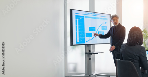 A middle-aged male business person giving an explanation in front of a large screen monitor
