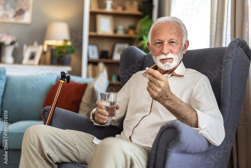 Sick senior man taking medication in nursing home. Senior man taking pills for depression sitting in armchair. Anxious senior patient swallowing pill with glass of water.