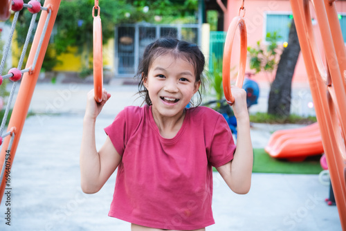 Asian cute young girl plays on the playground on a summer or spring day. Kid hanging on playground rings in park. Adorable child aged 9-10 years old.