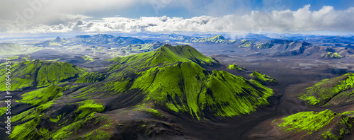 Εκτύπωση καμβά Aerial view of Iceland's rugged volcanic terrain with green moss covered mountains, dark craters, and valleys under a partly cloudy sky
