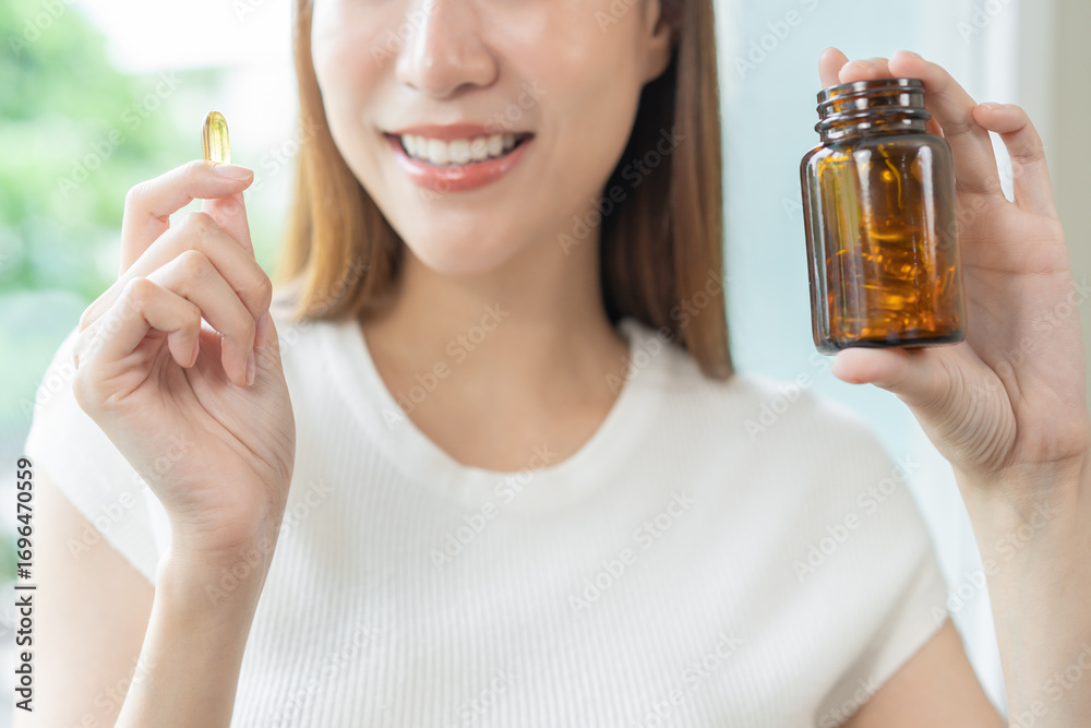 Fototapeta premium Dietary supplement concept, happy asian young woman hand holding fish oil pills, drugs medical tablet pouring capsules from medication bottle, take vitamin treatment for skin, hair at home, healthcare