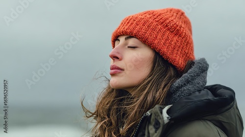 Portrait of young woman by the sea in knitted hat and winter jacket, eyes closed practicing mindful breathing, calm expression, serene moment of inner peace, relaxation and mindfulness