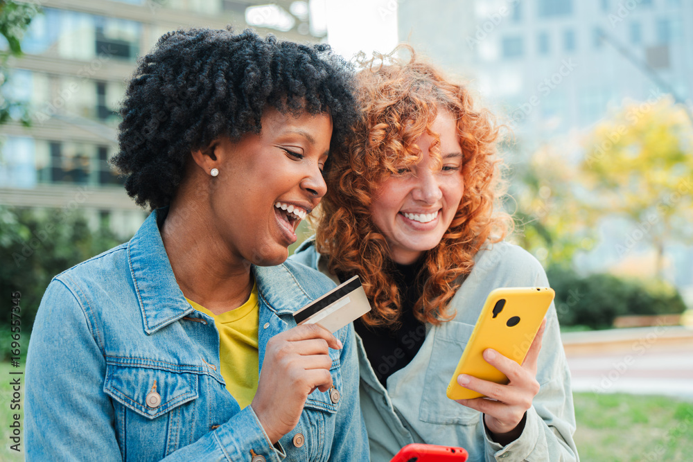 Fototapeta premium Two cheerful women happily engaging in a purchase using their smartphones and a credit card, showcasing the joy of shopping together in a lively manner.