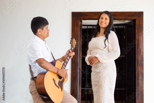 Novios enamorados en vestido blanco, abrazo romántico al aire libre con guitarra y espacio publicitario en pared blanca