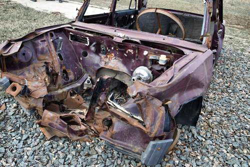 Rusted wreckage of abandoned car body on gravel patch.