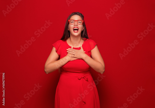 Thank you. Grateful young woman looking up with hand on his chest, isolated on red background. Rejoicing girl celebrates success.
