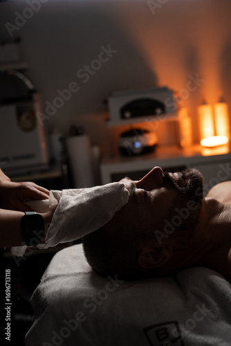 A man is lying on a massage table covered with a towel while receiving a facial spa treatment. A professional is gently applying a towel or compress to his face. The atmosphere is calm and relaxing