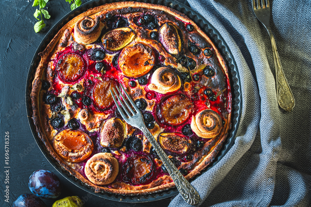 Fototapeta premium A tart or open pie with blueberries, figs, prunes and apricots in a baking dish on a gray background with a fork and napkin on the table. Top view. High quality photo