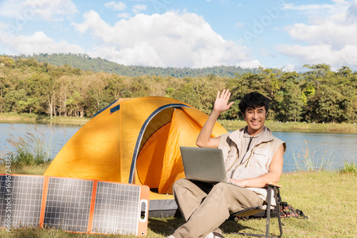 A young Asian man is happy working on his laptop notebook while traveling and camping beside a lake on sunny day connecting with solar cell energy