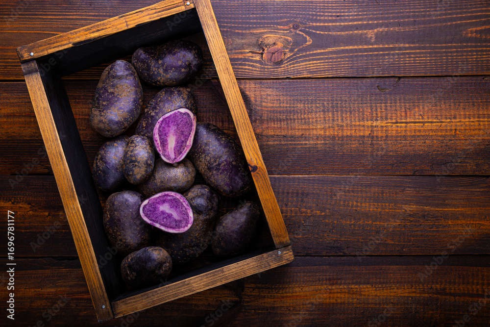 Obraz premium Harvest of ripe purple potatoes, unusual raw potatoes in the box on wooden background top view