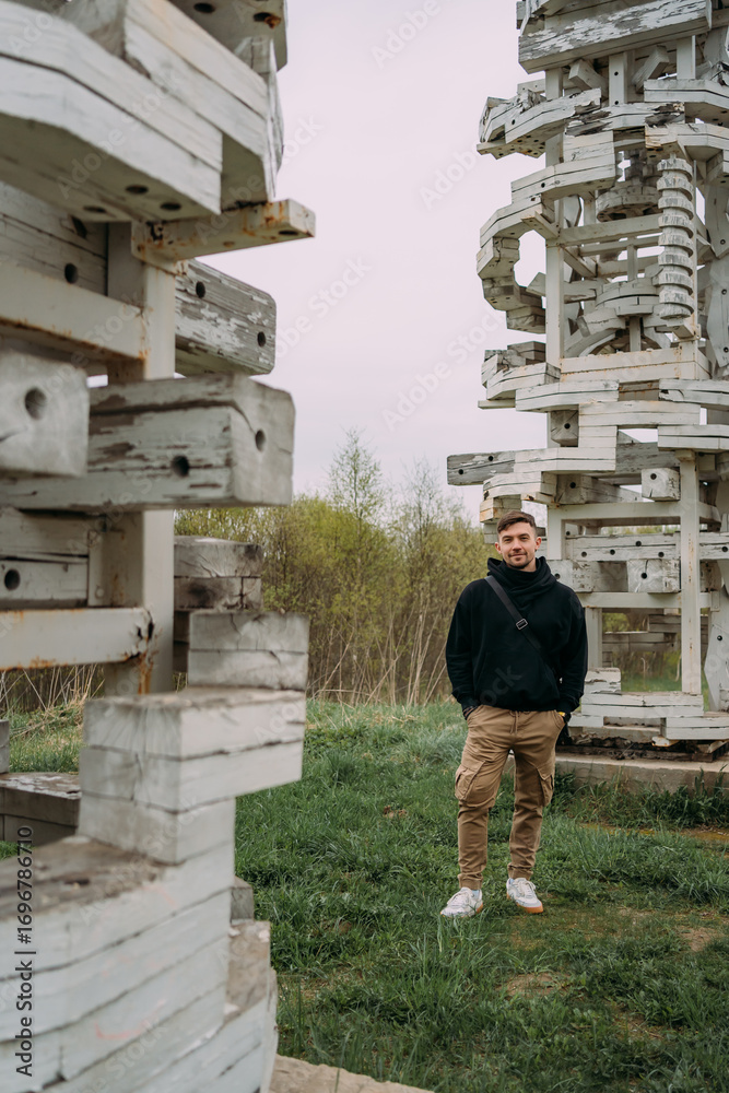 Fototapeta premium Young man standing between abstract white wooden sculptures in a grassy park under overcast sky