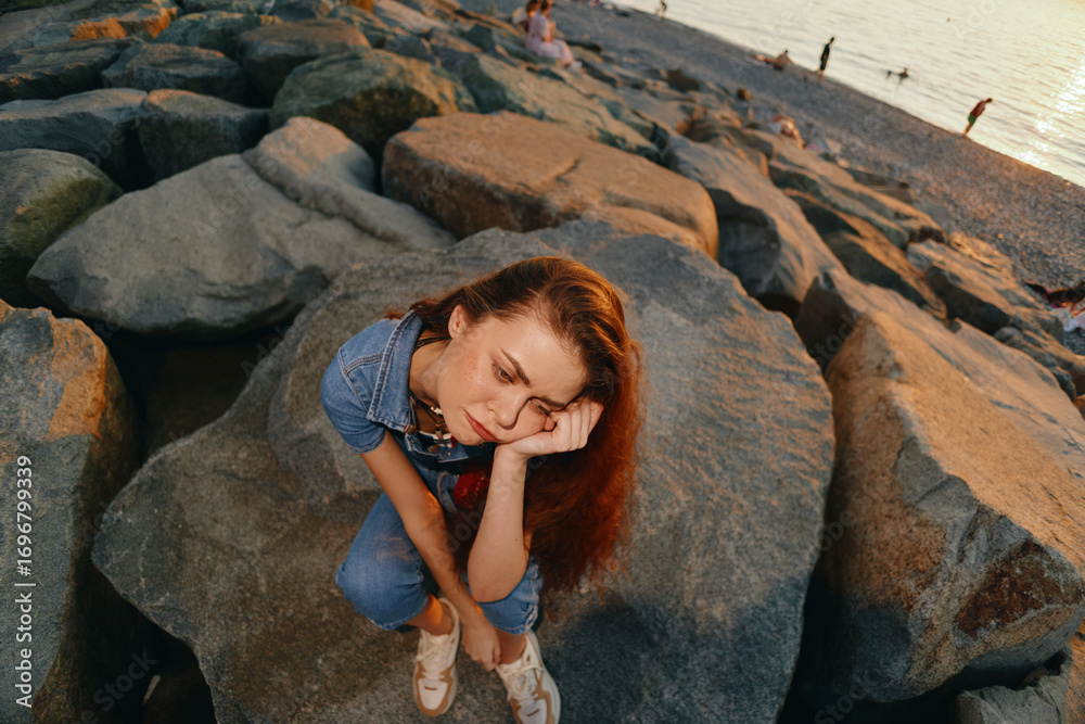 Fototapeta premium lifestyle woman in modern boho-western denim street style sitting on rocks at beach with warm film color tones, thoughtful mood, casual summer outfit